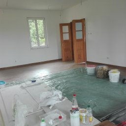 UKR-POL BUD Yurii Herchuk - Pokój w trakcie odświeżania: zabezpieczona folią podłoga, otwarte drewniane drzwi, puszki z farbą i akcesoria malarskie na blacie. Widok na okno z zielenią.