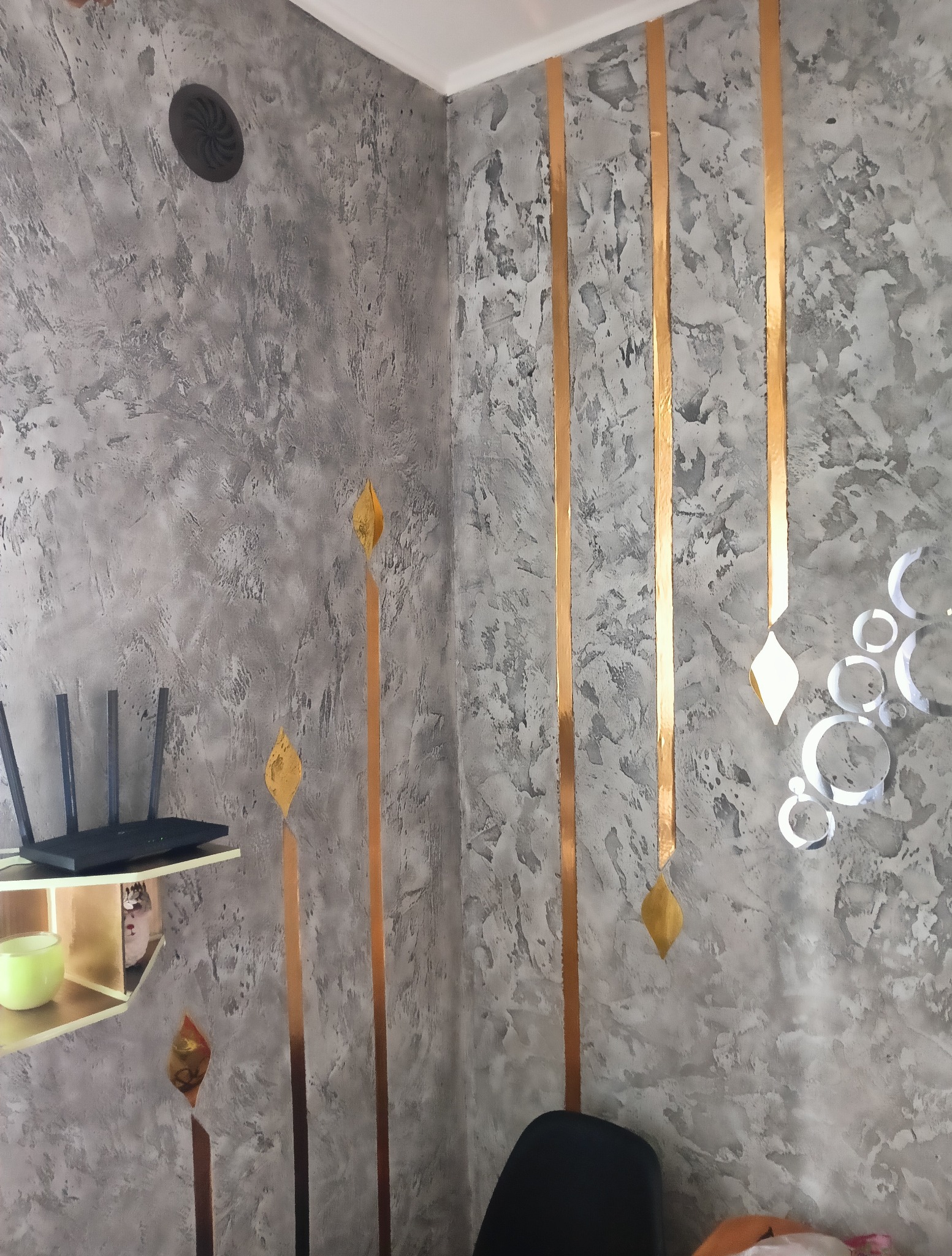 Dekoracyjna ściana w odcieniach szarości z pionowymi, złotymi pasami i ozdobami. Na ścianie półka z routerem i zielonym kubkiem. Widoczny fragment krzesła.