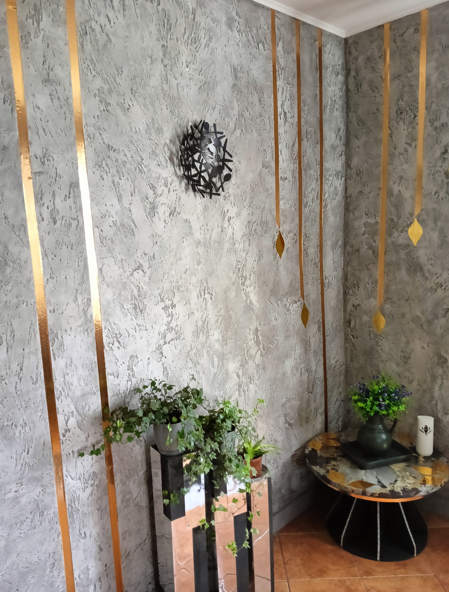 Wykończenie wnętrza: ściana z szarą strukturą, złote pasy dekoracyjne, nowoczesny kinkiet, rośliny doniczkowe na stoliku z lustrzanym blatem i okrągłym stoliku.