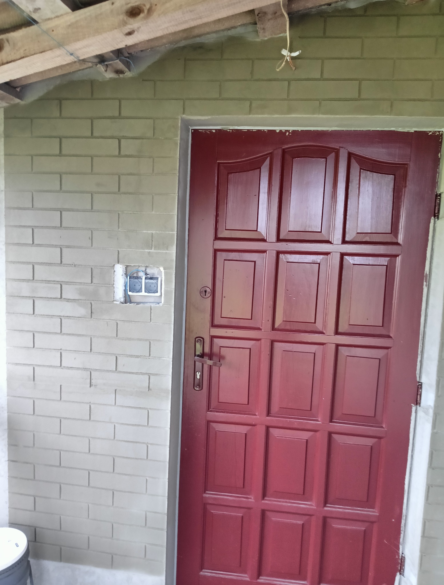 Ceglana ściana z gniazdkiem elektrycznym obok bordowych drzwi wejściowych. Widoczne drewniane elementy konstrukcyjne dachu.