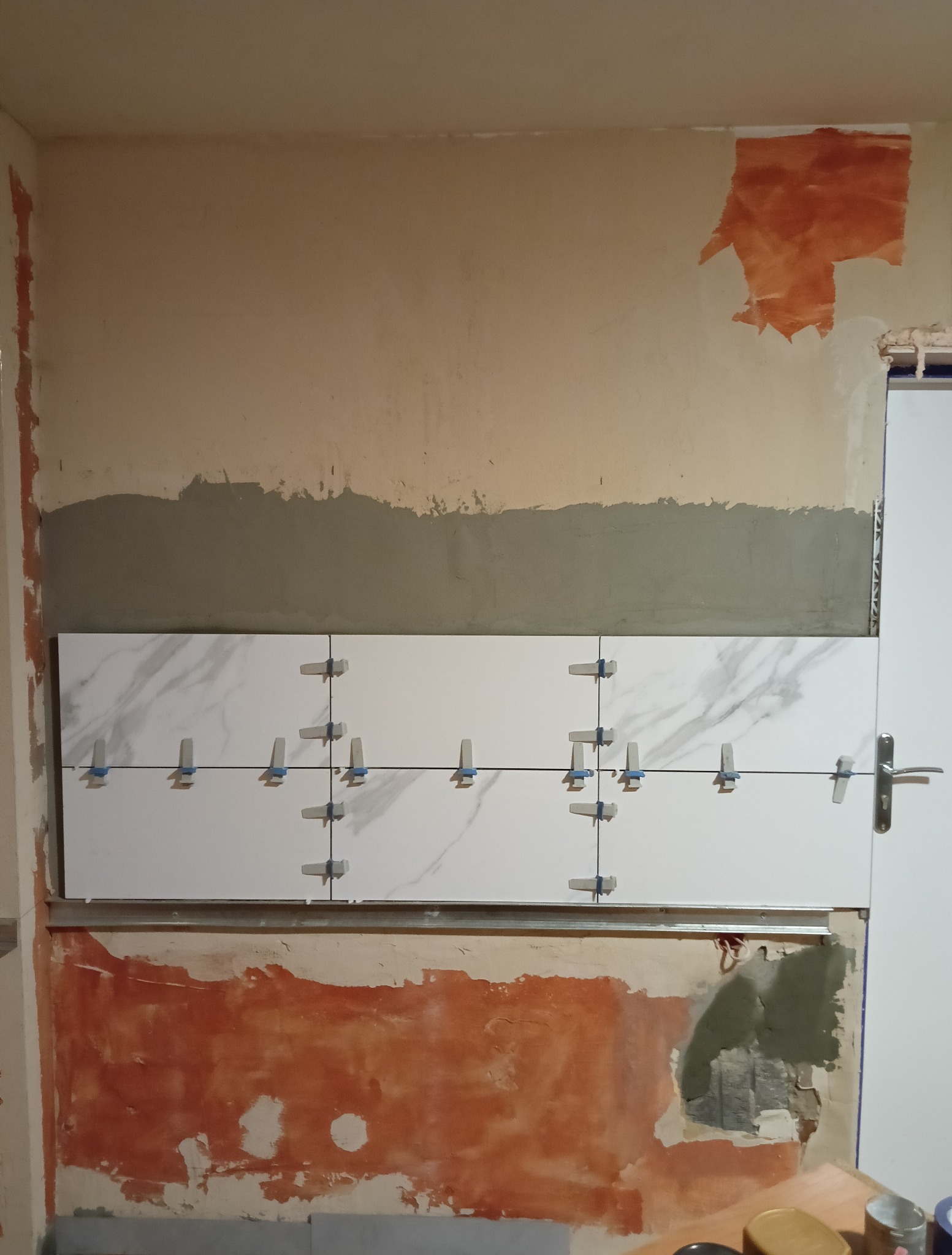 Układanie białych płytek imitujących marmur z użyciem krzyżyków dystansowych na ścianie w trakcie remontu w Bielany-Żylaki. Widoczne ślady po starej farbie.