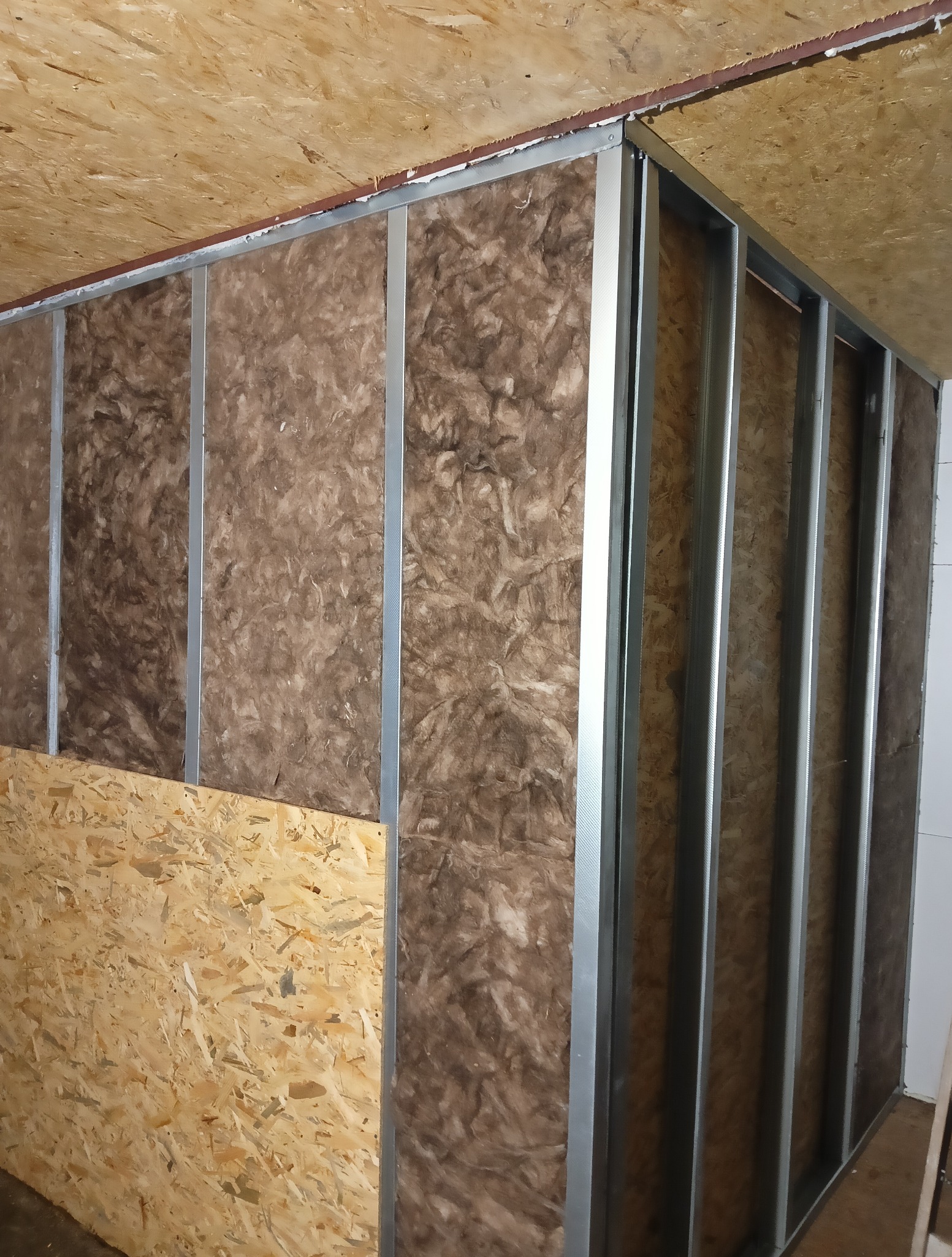 Szkielet ściany z metalowych profili wypełniony wełną mineralną, częściowo zakryty płytą OSB. Widoczne elementy konstrukcyjne i izolacyjne.