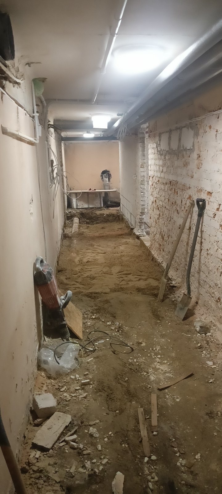Wnętrze w trakcie prac przygotowawczych: odsłonięta cegła, ziemia, narzędzia (szpadle, młot pneumatyczny). Widok na korytarz z surowym wykończeniem i widocznymi instalacjami.