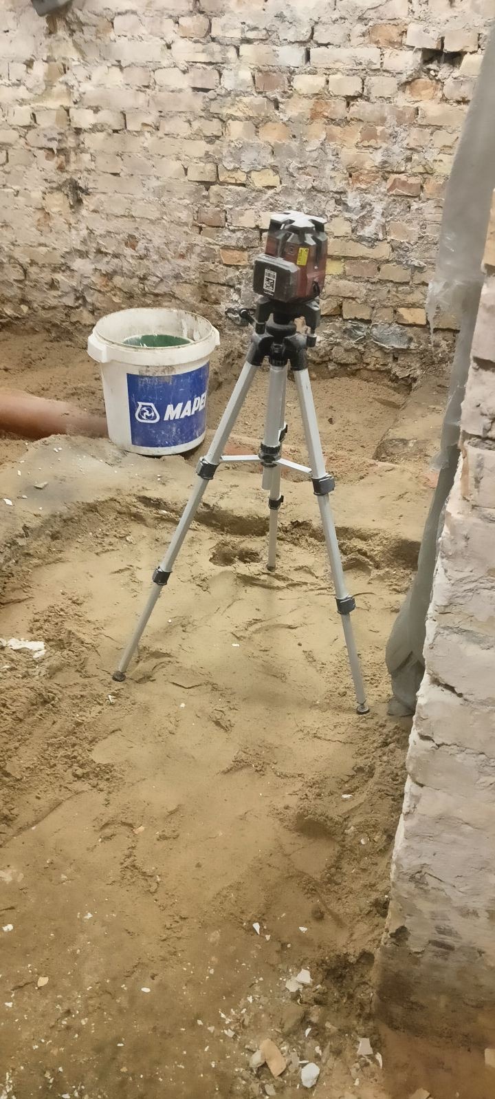 Laser krzyżowy na statywie, ustawiony na piasku w pomieszczeniu z ceglanymi ścianami. Obok stoi białe wiadro z logo Mapei. Przygotowanie do prac niwelacyjnych.