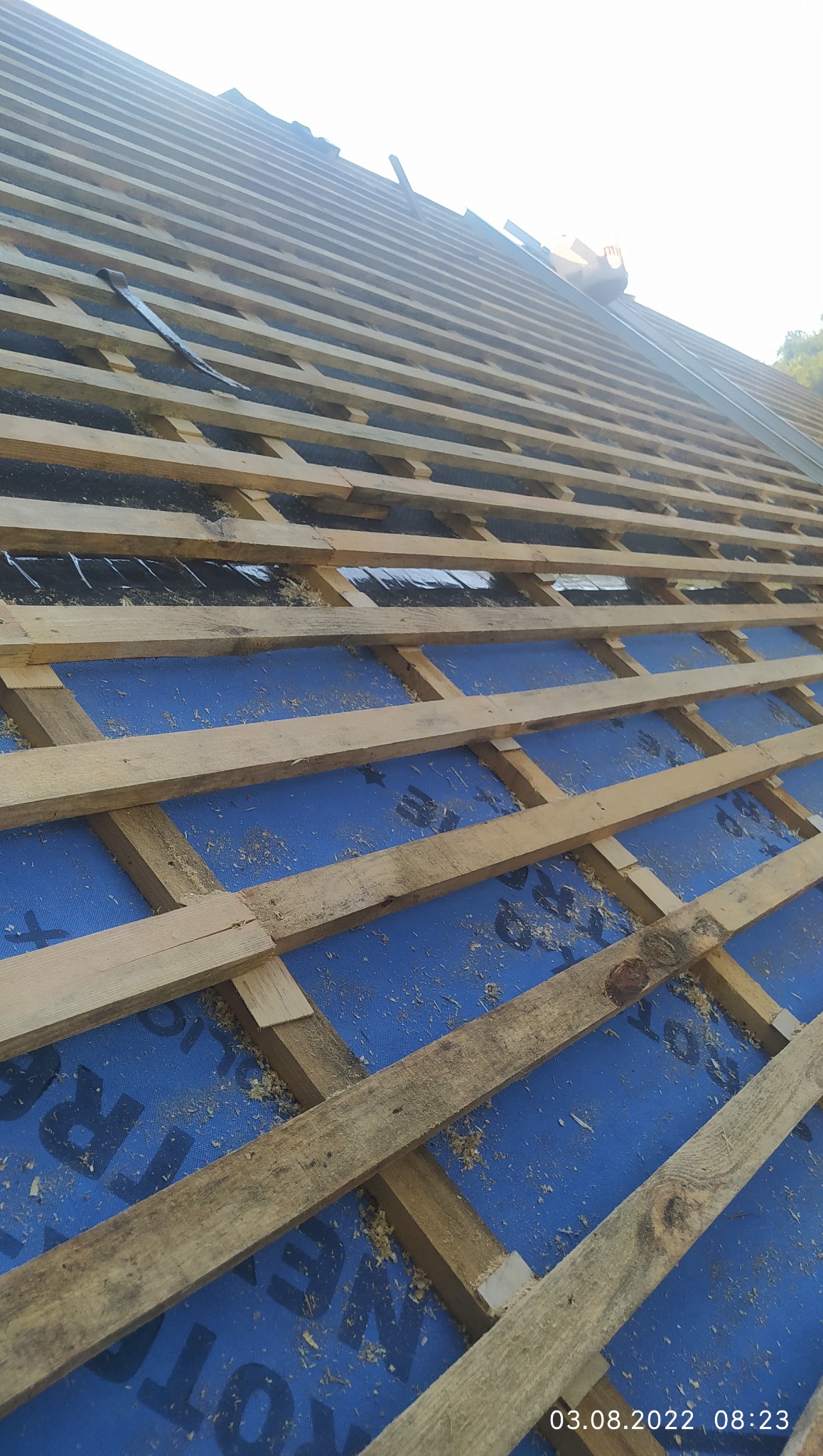 Konstrukcja dachu z drewnianych łat na niebieskiej membranie, widok z dołu pod kątem. Elementy mocowania widoczne. Jasne światło słoneczne.