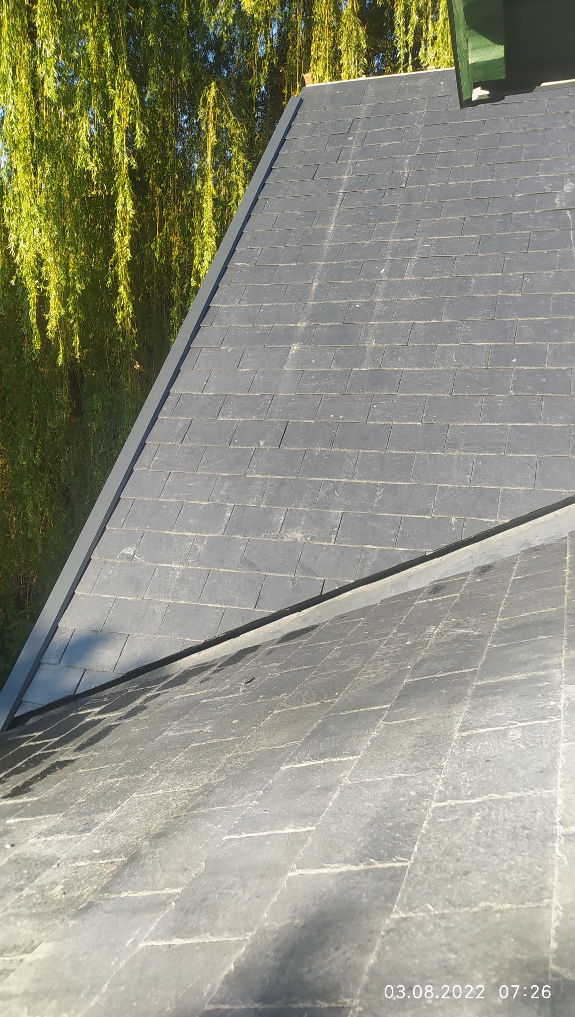 Szary dach z łupka w promieniach słońca, z widocznym fragmentem rynny i zielenią drzew w tle. Układanie dachówki w regularny wzór, widoczne detale faktury materiału.