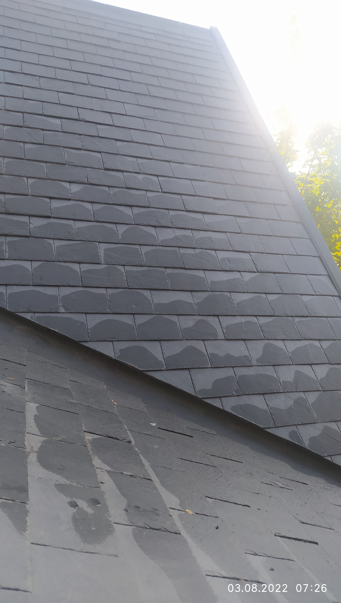 Szary dach pokryty dachówką bitumiczną w układzie łuskowym, lekko mokry od deszczu, z widocznym fragmentem drzewa w tle i datą wykonania zdjęcia w prawym dolnym rogu.