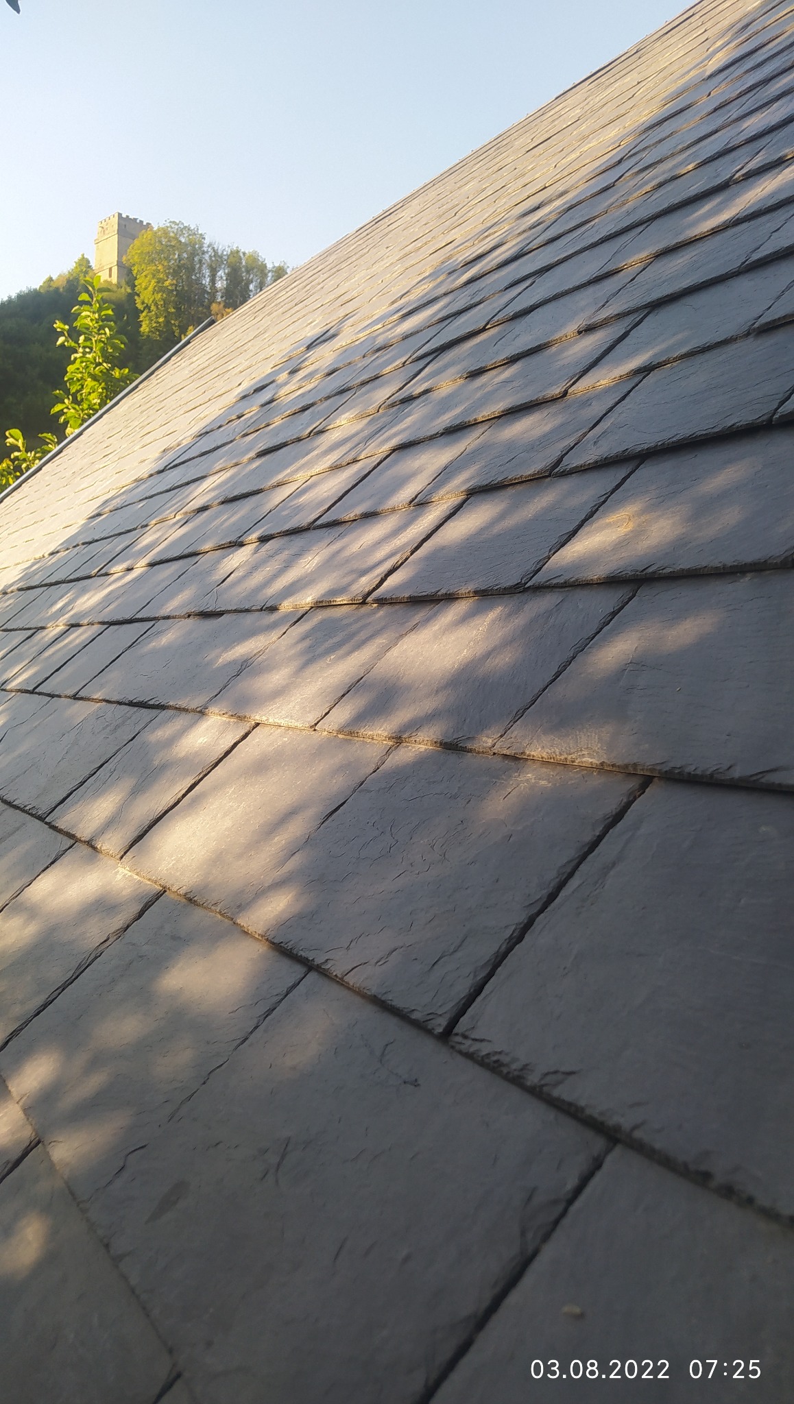 Szary dach pokryty łupkiem w promieniach słońca, z widocznym zamkiem w tle. Ujęcie z dołu podkreśla teksturę i regularność ułożenia płyt.