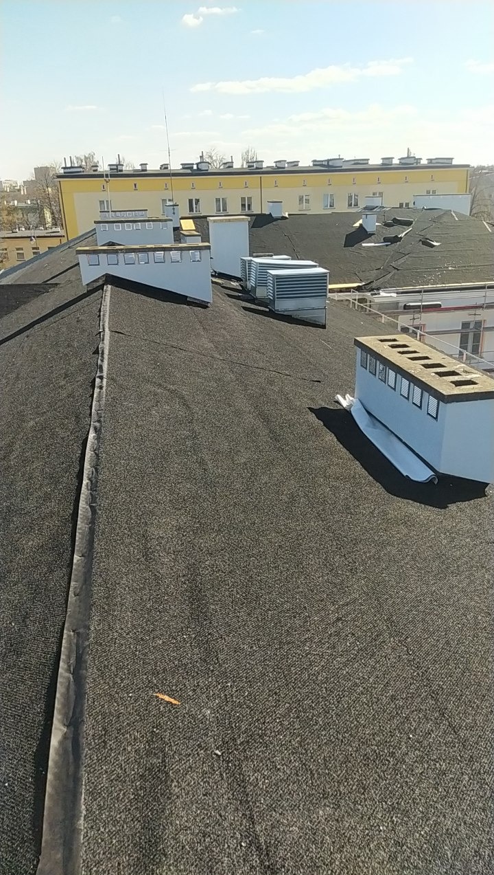 Czarny dach pokryty papą termozgrzewalną z białymi elementami wentylacyjnymi i klimatyzacyjnymi na tle budynku w słoneczny dzień. Widok z góry.