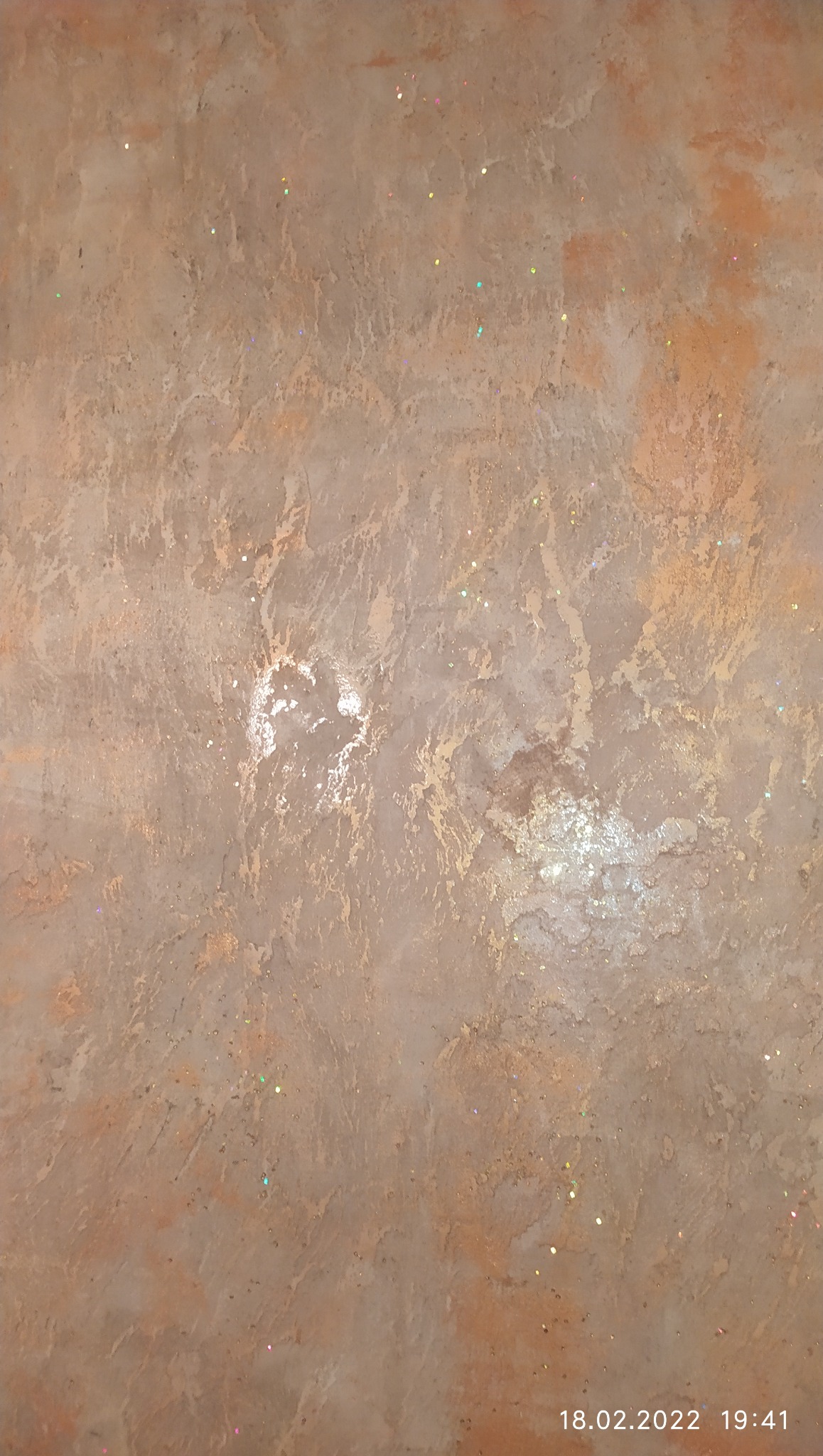 Dekoracyjna ściana w odcieniach beżu i brązu z efektem przetarć i delikatnymi, kolorowymi drobinkami brokatu, tworząca unikalną teksturę. Widoczne refleksy światła.