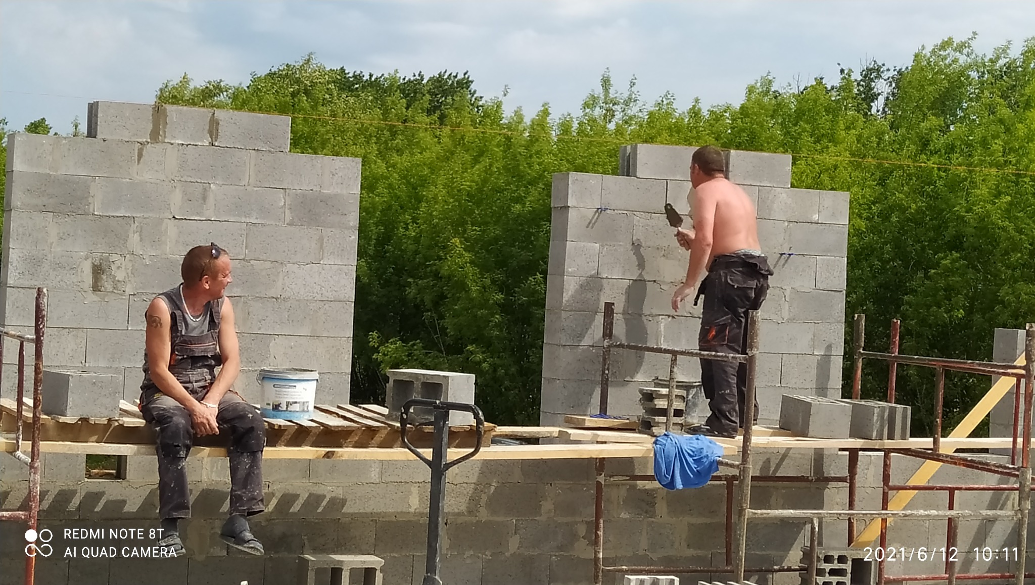 Wznoszenie ścian z bloczków betonowych na budowie domu jednorodzinnego. Dwóch robotników na rusztowaniu, jeden muruje, drugi odpoczywa. Widok na zieleń w tle.