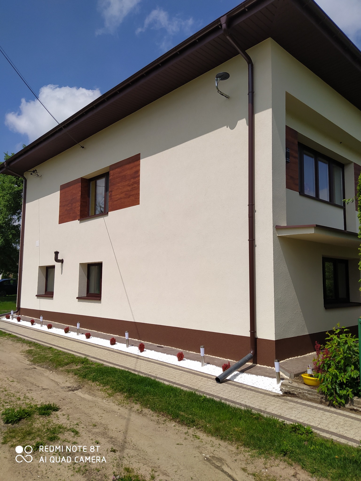 Elewacja dwukondygnacyjnego domu z beżowym tynkiem, brązowymi akcentami i oknami. Widoczne rynny, oświetlenie zewnętrzne i dekoracyjny pas z białego kamienia.