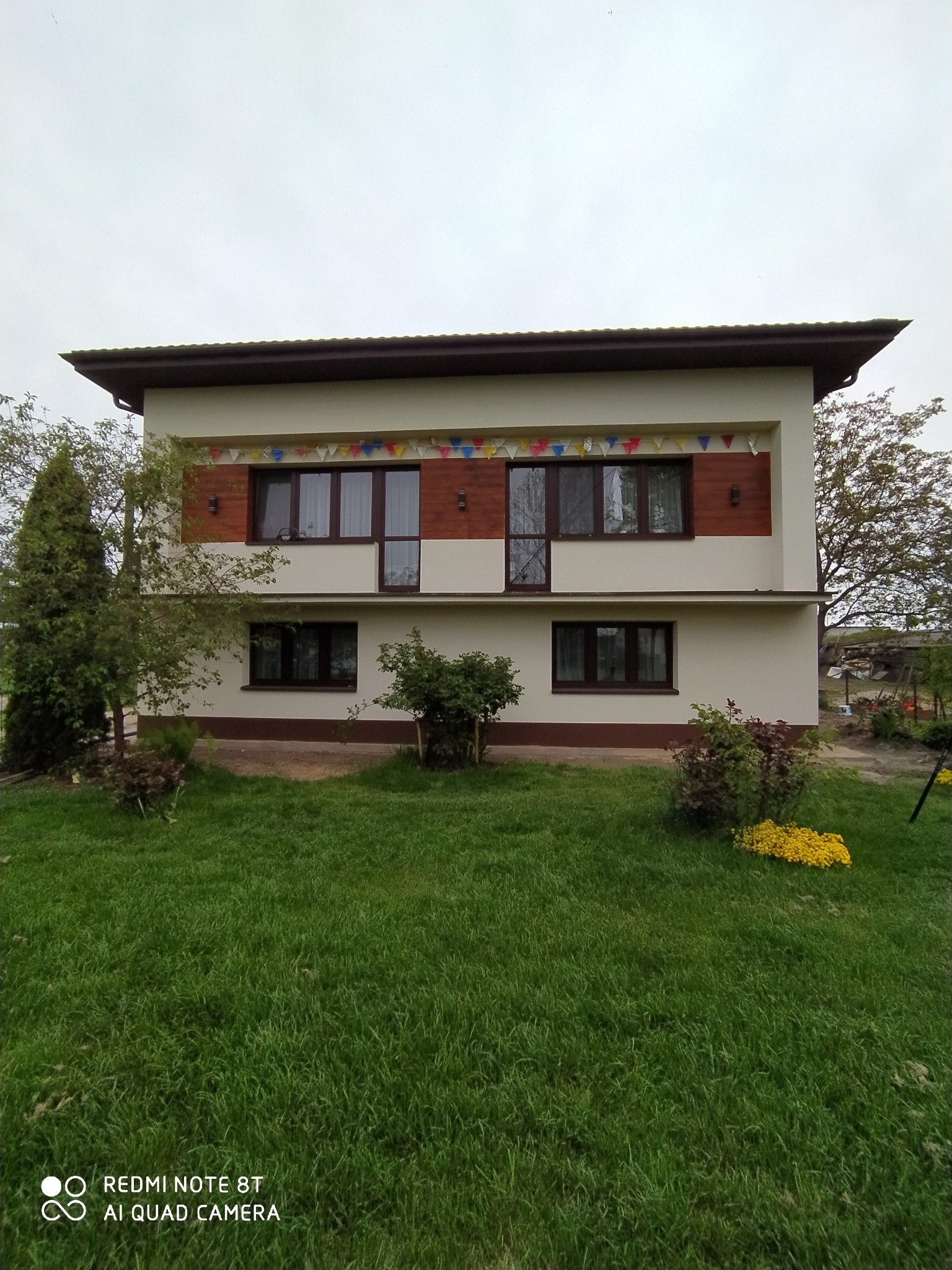 Dwupiętrowy dom z beżową elewacją, brązowymi oknami i dekoracyjnymi flagami. Widok z trawnika, drzewa w tle. Ujęcie z dołu, pochmurne niebo.