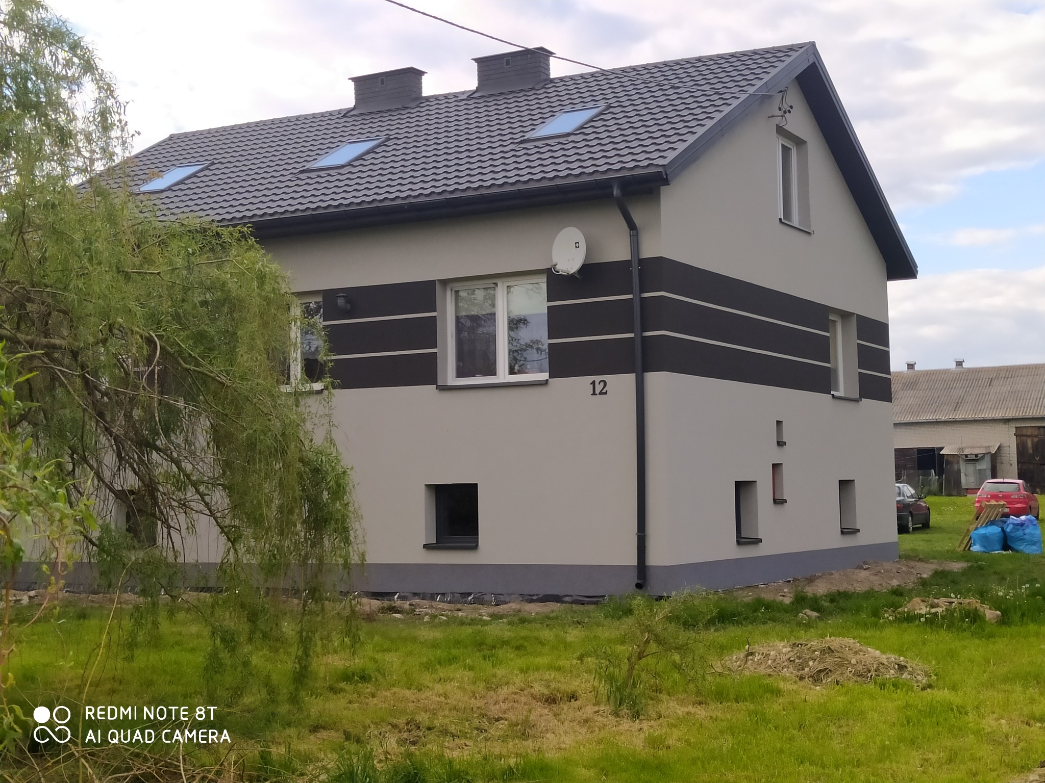 Odnowiony dom z szarą elewacją i ciemnymi pasami, widoczne okna dachowe i numer '12'. W tle budynki gospodarcze i drzewa. Ujęcie z poziomu trawnika.