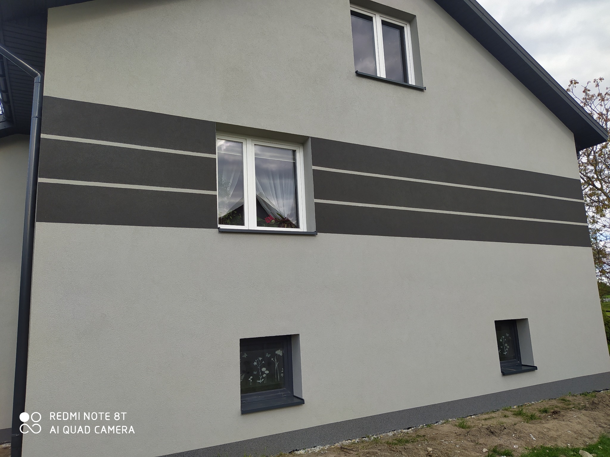 Elewacja domu w odcieniach szarości z poziomymi pasami. Widoczne okna z białymi ramami i fragment dachu. Świeżo pomalowana fasada budynku.
