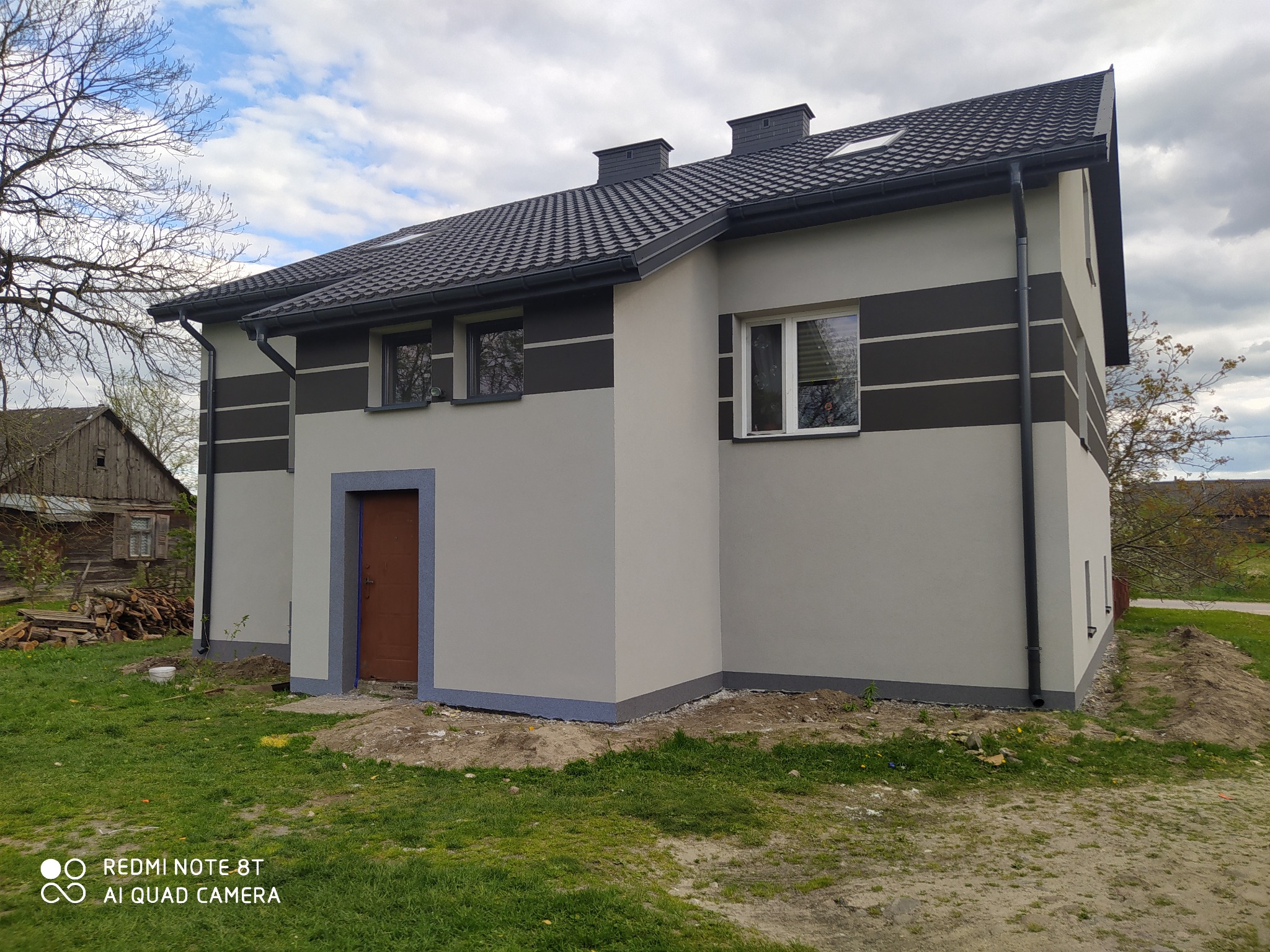 Nowoczesny dom z szarą elewacją i ciemnymi pasami, brązowe drzwi wejściowe, czarny dach z dachówki. Widoczne okna z odbiciem drzew. Trawa i fragment starego budynku w tle.