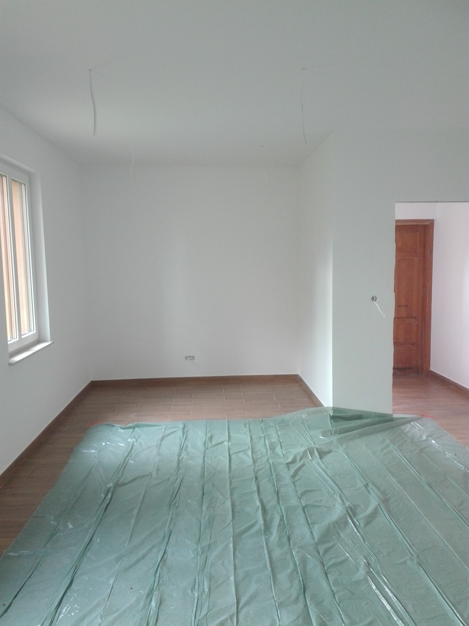 Białe ściany w pokoju po remoncie, zabezpieczone folią malarską. Widoczne nowe panele podłogowe i okno. W tle drewniane drzwi.