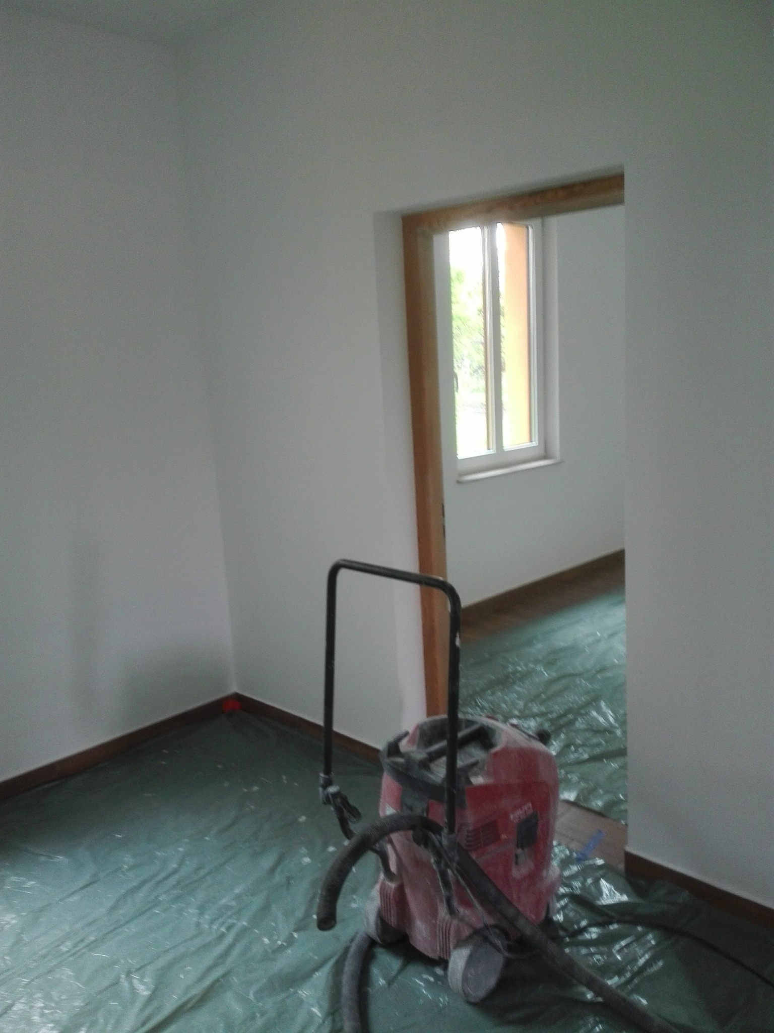 Przygotowanie do malowania: Pokój zabezpieczony folią malarską, widoczny odkurzacz przemysłowy Hilti. Świeżo pomalowane ściany na biało, drewniane listwy przypodłogowe.