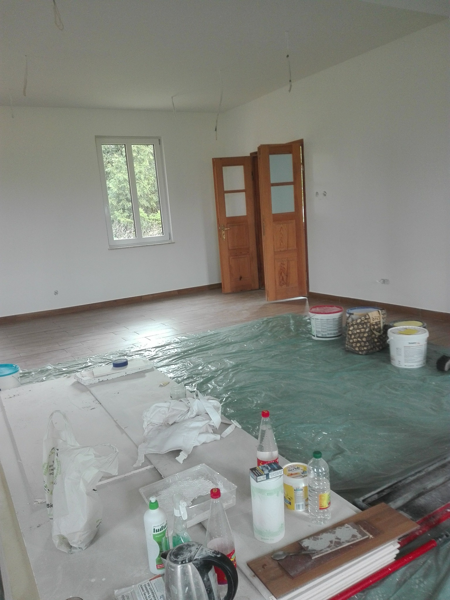 Pokój w trakcie odświeżania: zabezpieczona folią podłoga, otwarte drewniane drzwi, puszki z farbą i akcesoria malarskie na blacie. Widok na okno z zielenią.