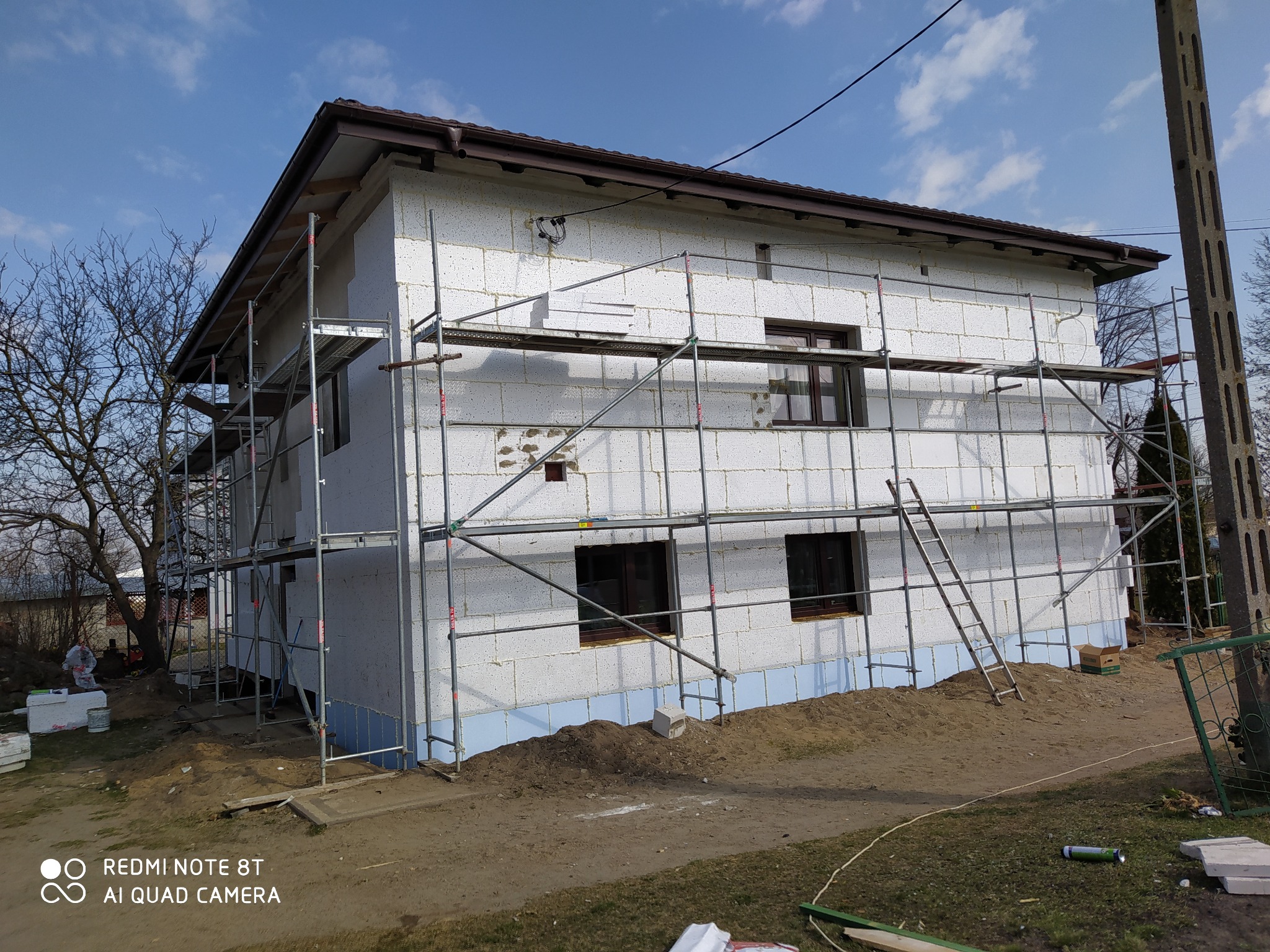 Budowa domu w trakcie ocieplania ścian zewnętrznych. Widoczne rusztowania, białe bloczki i niebieski styropian przy fundamentach. Częściowo zamontowane okna.