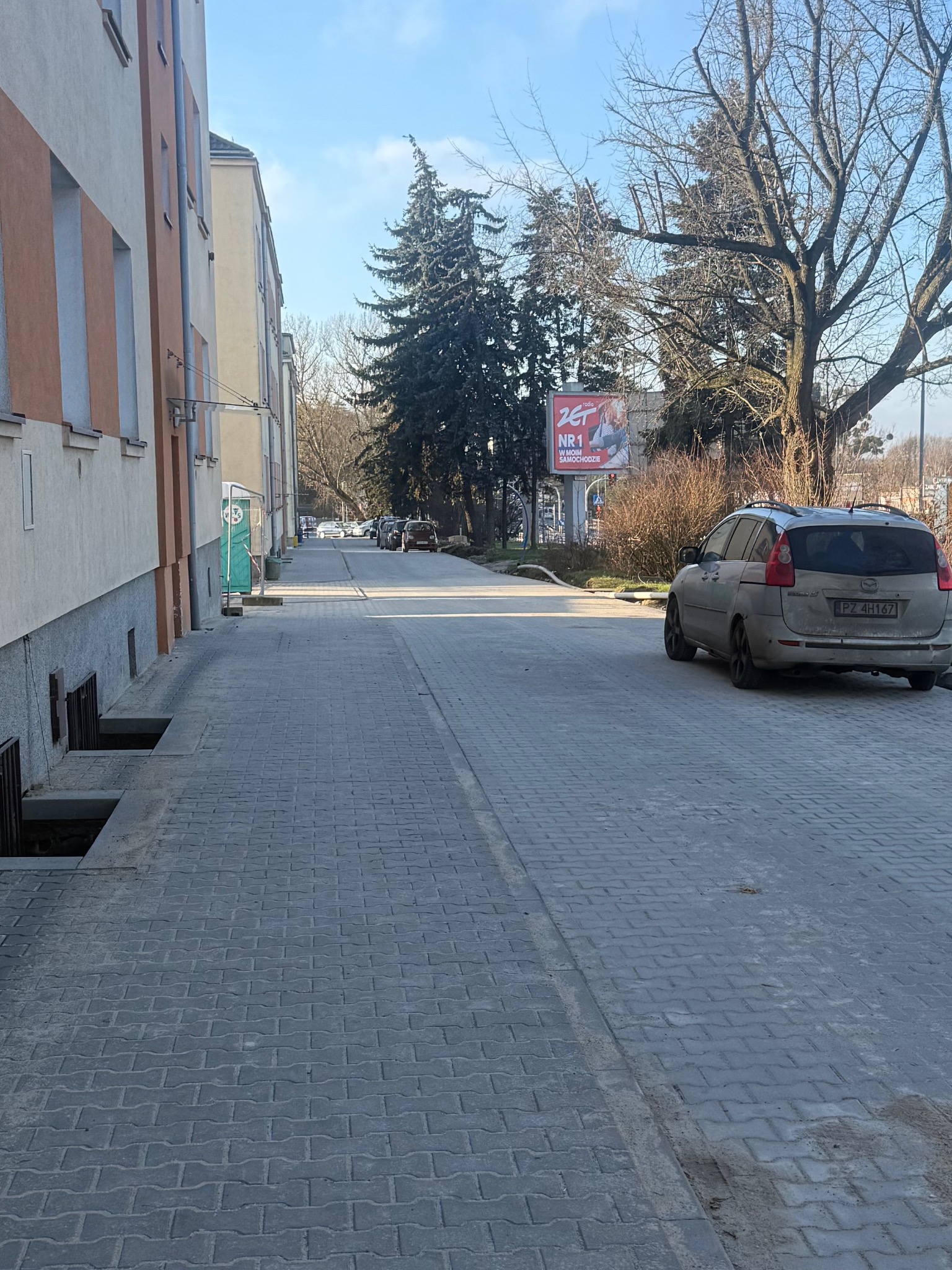 Ułożona szara kostka brukowa na długim podjeździe między budynkami, z zaparkowanym samochodem i widocznym billboardem w tle.