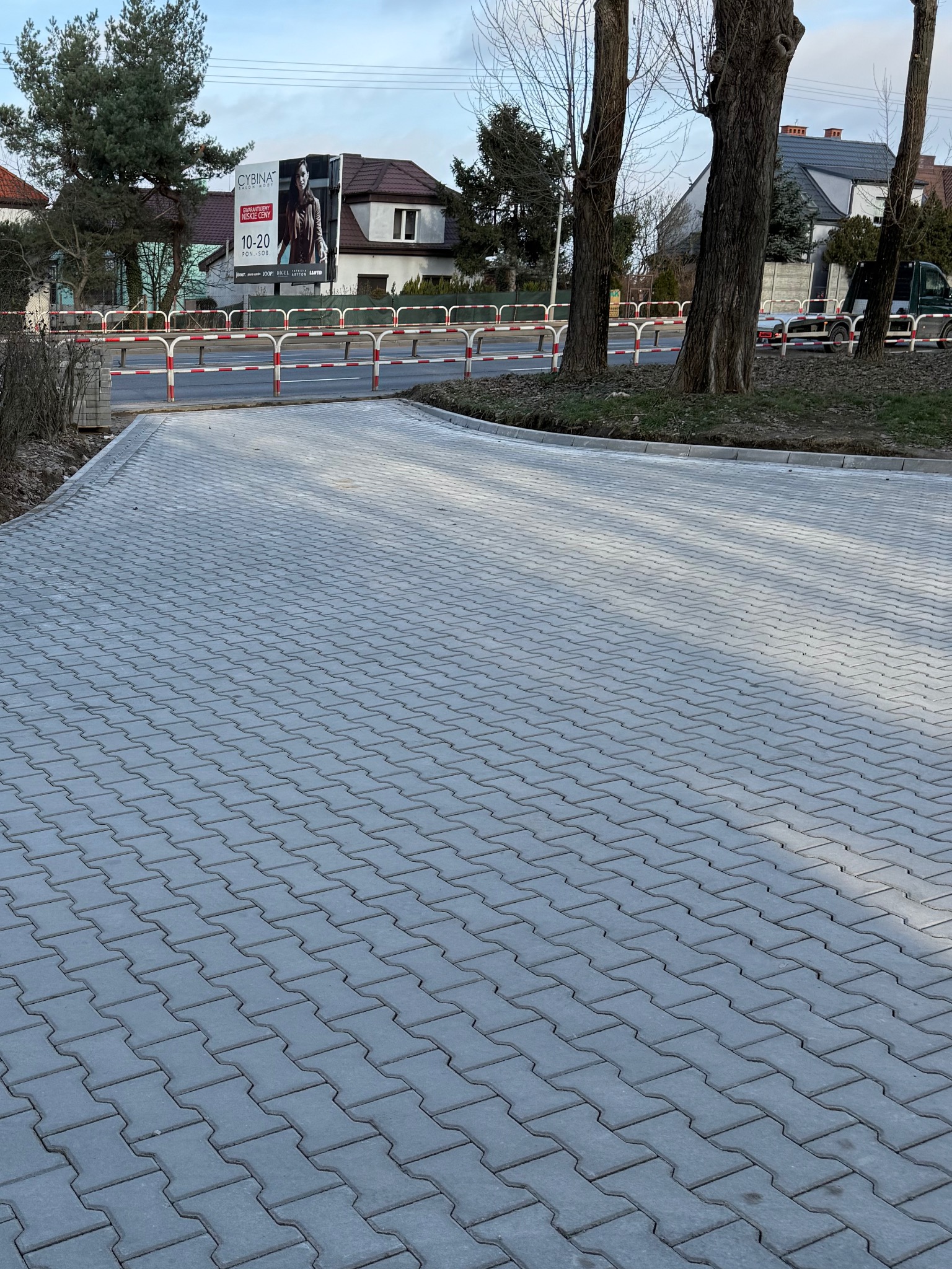 Szara kostka brukowa ułożona na podjeździe, widok z góry, w tle drzewa i budynki. Nowa nawierzchnia z kostki, równo ułożona, z widocznymi liniami podziału.