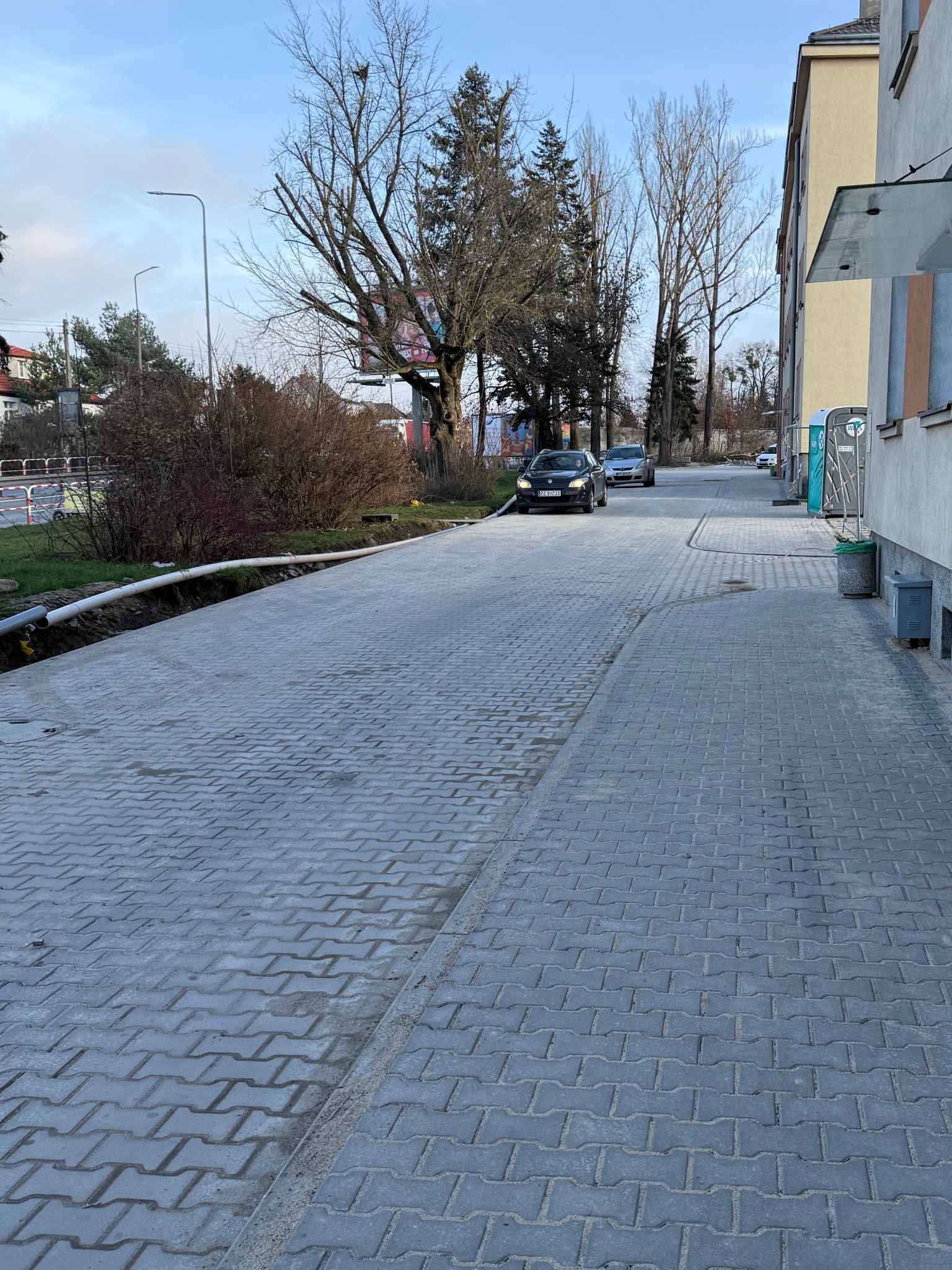 Ułożona szara kostka brukowa na chodniku miejskim, z widocznymi elementami infrastruktury i zieleni w tle. Widoczne samochody na ulicy w oddali.
