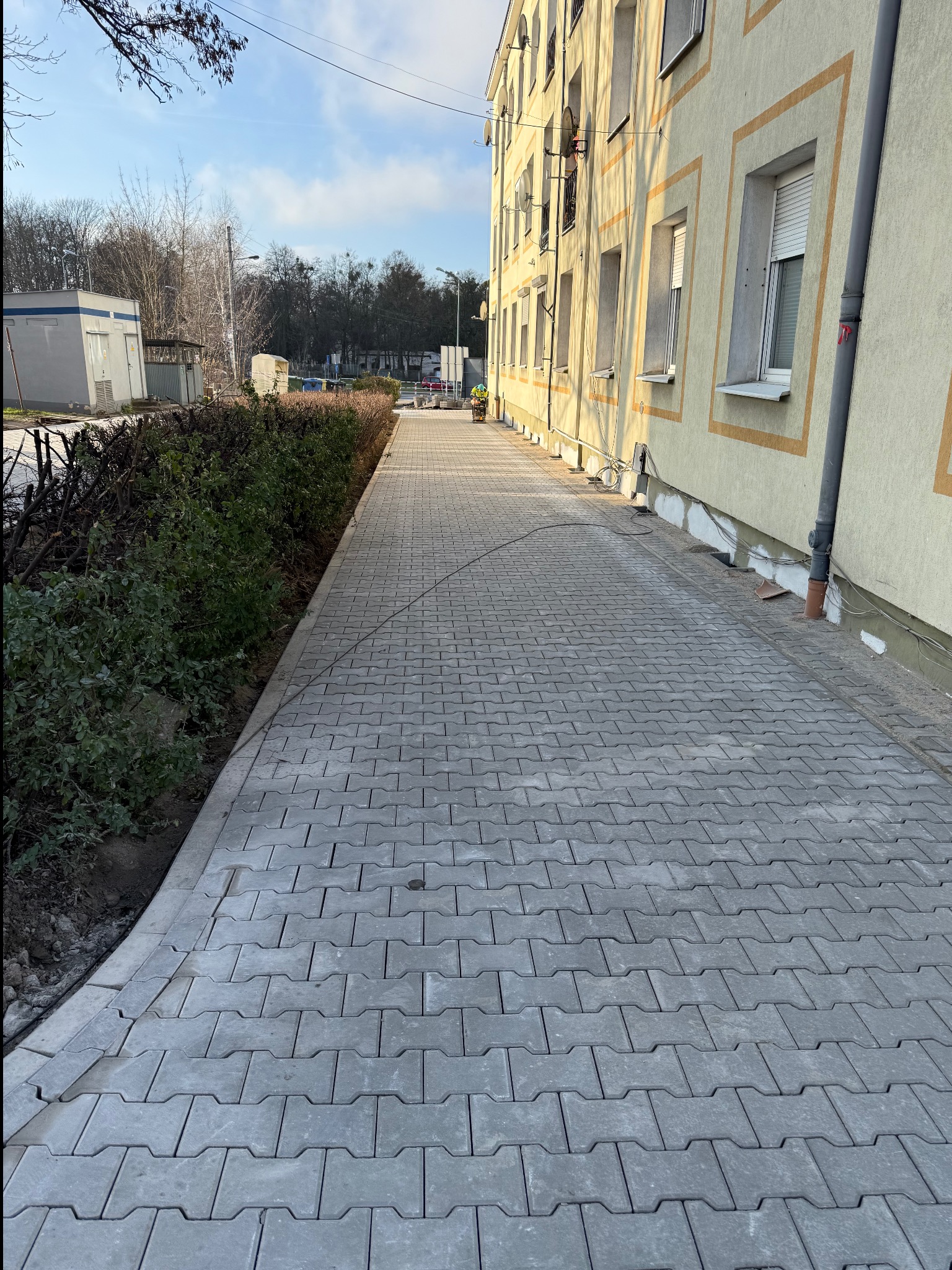 Szara kostka brukowa ułożona wzdłuż budynku z żółtymi obramowaniami okien, obok przycięte krzewy. Widoczny fragment instalacji na elewacji.
