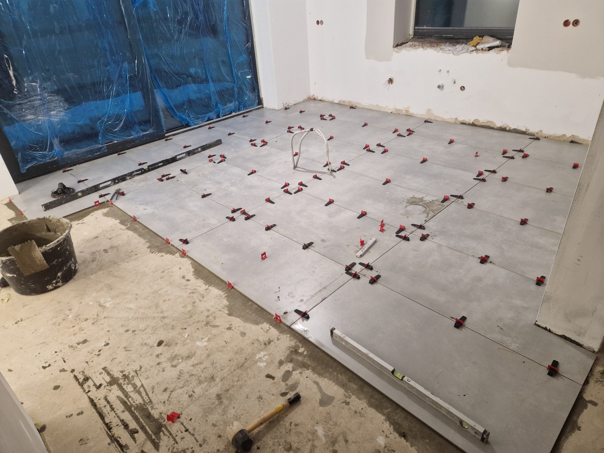 Układanie szarych płytek podłogowych z użyciem poziomicy i czerwonych klipsów poziomujących. Widoczny kubeł i młotek gumowy. Okno w stanie surowym.