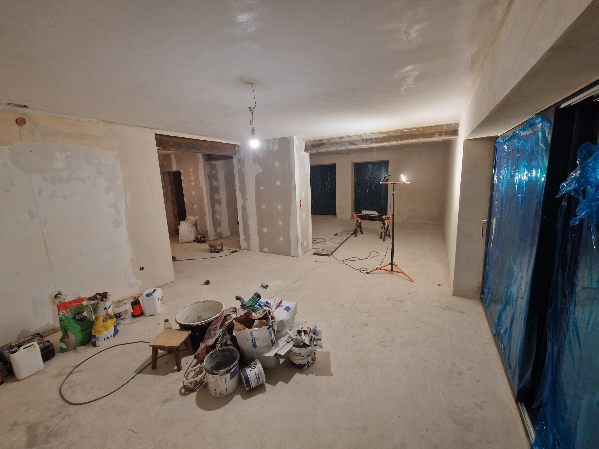 Wnętrze w trakcie remontu: surowy beton, ściany przygotowane do szpachlowania, narzędzia i materiały budowlane, okna zabezpieczone folią. Widoczny etap prac wykończeniowych.