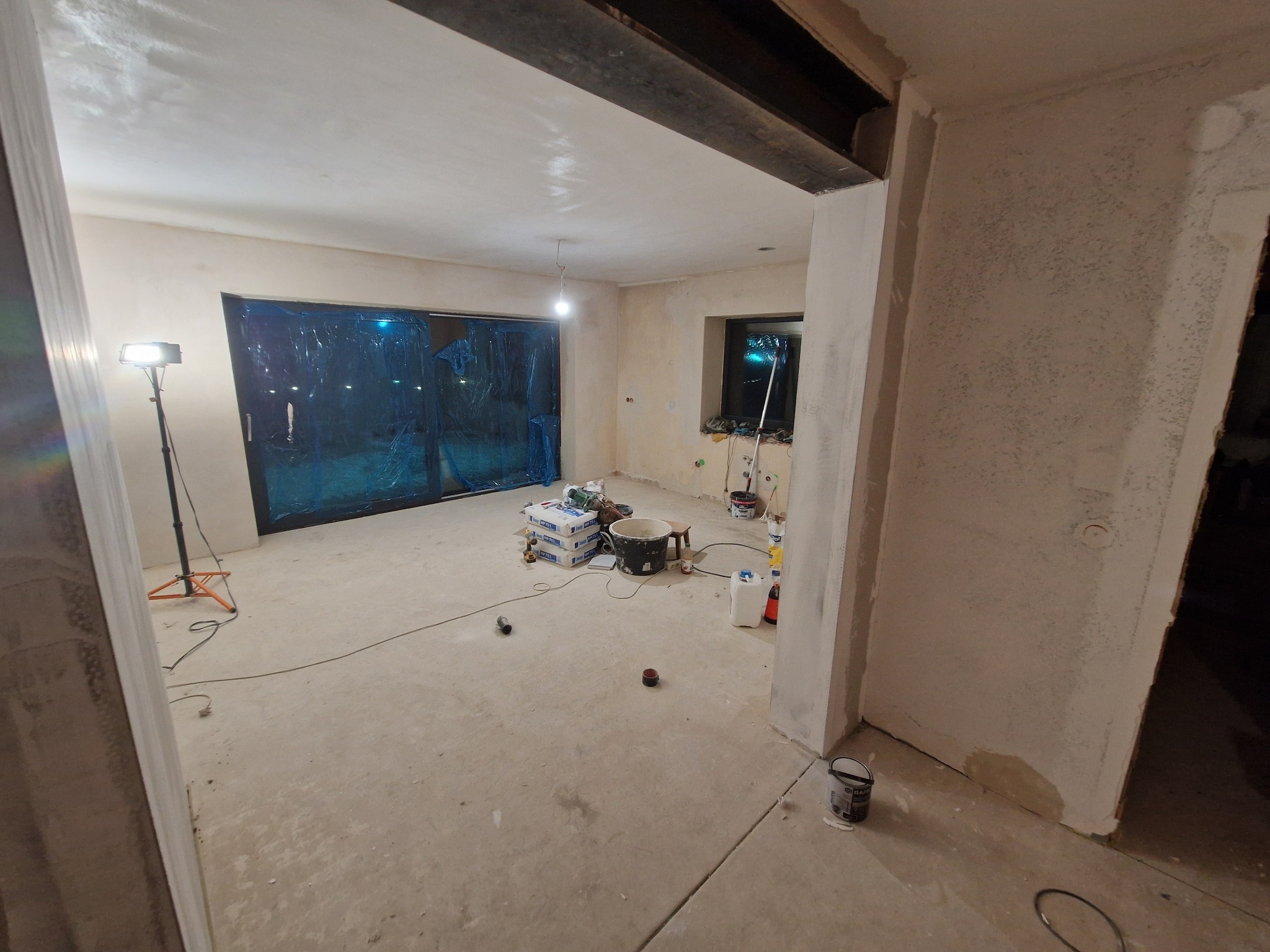 Wnętrze w trakcie szpachlowania: ściany pokryte gładzią, okna zabezpieczone folią, widoczne narzędzia i materiały budowlane, lampa oświetlająca pomieszczenie.