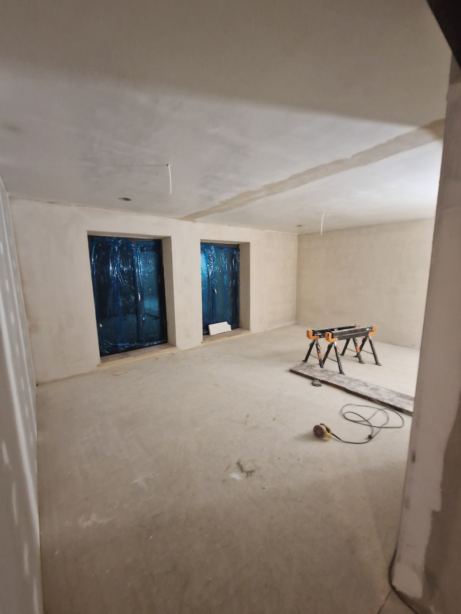 Wnętrze w trakcie szpachlowania: ściany pokryte gładzią, okna zabezpieczone folią, widoczne kozły i szlifierka. Etap przygotowania do dalszych prac wykończeniowych.