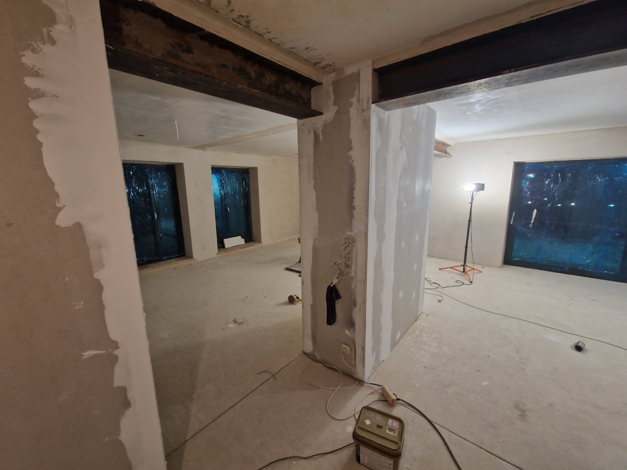 Wnętrze w trakcie szpachlowania ścian i sufitu, widoczne stalowe belki, okna zabezpieczone folią, podłoga betonowa, lampa budowlana. Prace wykończeniowe w surowym stanie.