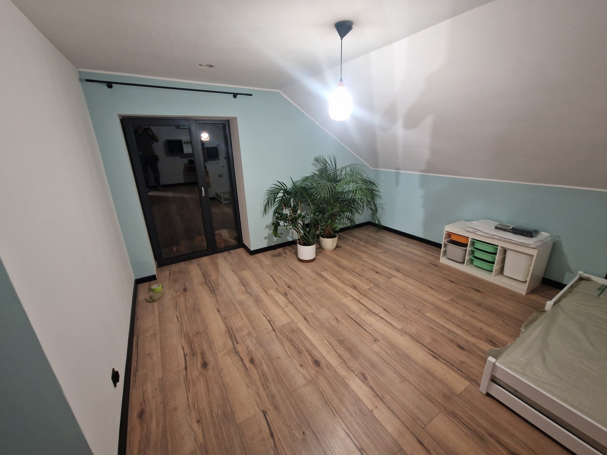 Jasne wnętrze poddasza z drewnianą podłogą, roślinami doniczkowymi i meblami. Ściany w odcieniach bieli i błękitu. Widoczne drzwi balkonowe z czarną ramą.