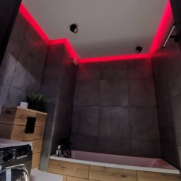 DS Wykończenia Dariusz Szczygłowski - Nowoczesna łazienka z szarymi płytkami, drewnopodobną zabudową wanny i pralki. Czerwone oświetlenie LED pod sufitem dodaje wnętrzu charakteru. Minimalistyczne lampy sufitowe.