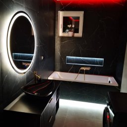 DS Wykończenia Dariusz Szczygłowski - Nowoczesna łazienka w czerni z marmurowym wzorem, podświetleniem LED i owalnym lustrem. Czarna umywalka na szafce z ryflowanymi frontami, biała wanna z złotą armaturą.