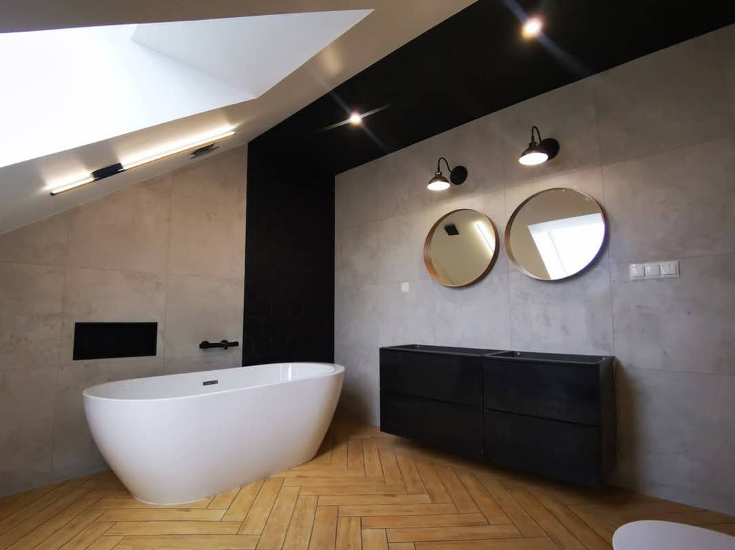 Nowoczesna łazienka na poddaszu z wolnostojącą wanną, drewnianą podłogą w jodełkę i czarnymi akcentami. Dwa okrągłe lustra nad czarną szafką.