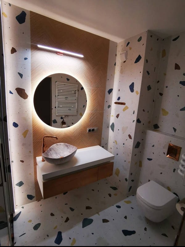 Nowoczesna łazienka z okrągłym lustrem LED, umywalką w kształcie muszli i wzorem lastryko na ścianach i podłodze. Minimalistyczny design z akcentami miedzianymi.