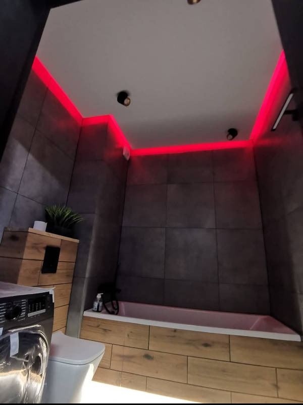 Nowoczesna łazienka z szarymi płytkami, drewnopodobną zabudową wanny i pralki. Czerwone oświetlenie LED pod sufitem dodaje wnętrzu charakteru. Minimalistyczne lampy sufitowe.
