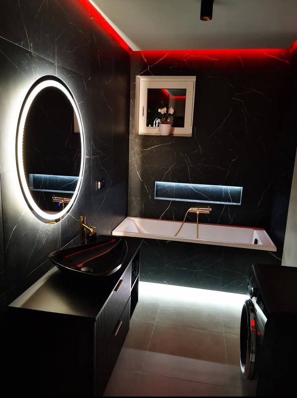 Nowoczesna łazienka w czerni z marmurowym wzorem, podświetleniem LED i owalnym lustrem. Czarna umywalka na szafce z ryflowanymi frontami, biała wanna z złotą armaturą.