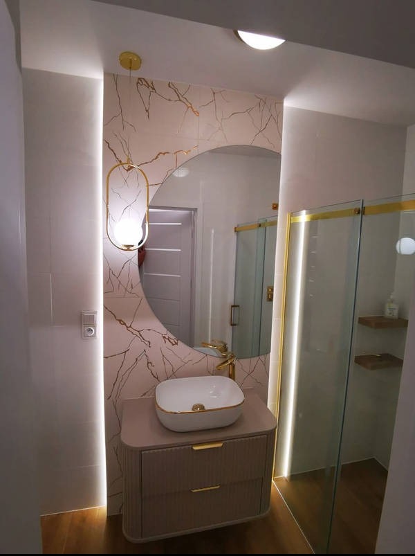 Elegancka łazienka z okrągłym lustrem, umywalką nablatową i kabiną prysznicową ze złotymi akcentami. Płytki imitujące marmur z żyłami. Oświetlenie LED.