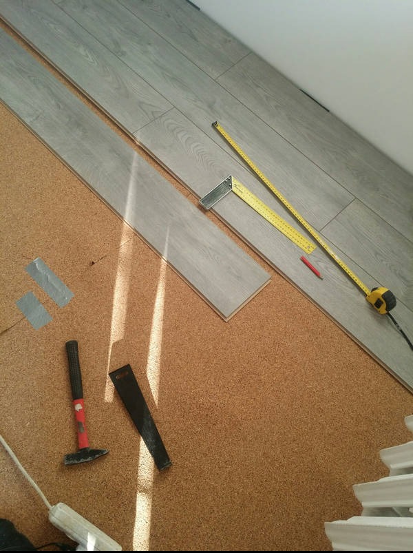 Układanie paneli podłogowych: widok z góry na panele w odcieniach szarości, narzędzia (miarka, kątownik, młotek) i podkład korkowy. Etap prac, światło słoneczne.