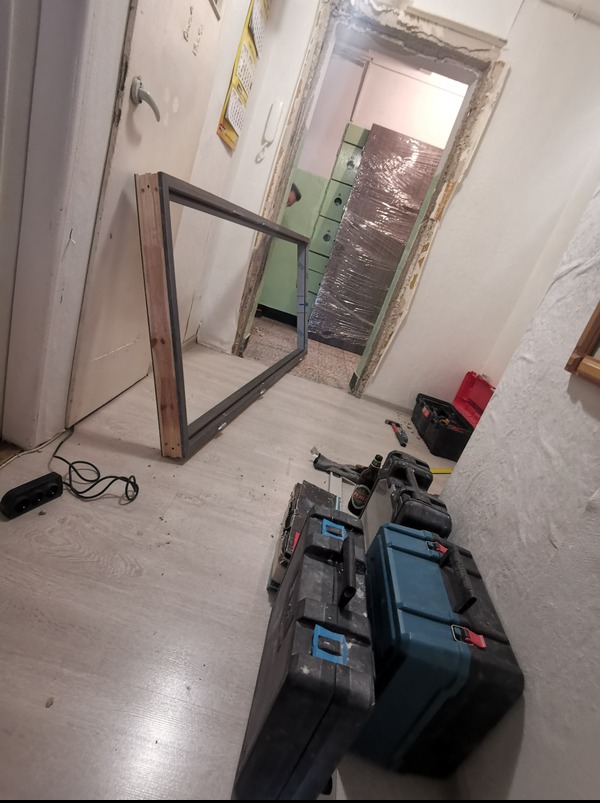 Przygotowanie do montażu ościeżnicy w remontowanym mieszkaniu. Widoczne narzędzia, skrzynki narzędziowe i zabezpieczone drzwi w folii. Prace wykończeniowe w toku.