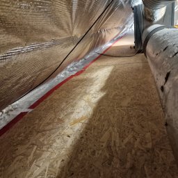 Vasyl Dovhanych - Skośny widok na poddasze z folią izolacyjną z czerwoną taśmą, drewnianą podłogą i fragmentem rury wentylacyjnej. Widoczne kable i instalacje.