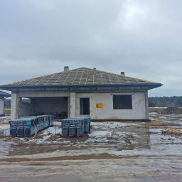 Dom w stanie surowym otwartym z widoczną konstrukcją dachu i stosem dachówek CREATON. Na pierwszym planie błoto i resztki śniegu. Pochmurne niebo.