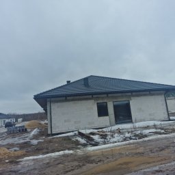 Vasyl Dovhanych - Nowoczesny dom jednorodzinny w trakcie budowy, z ciemną dachówką i białymi ścianami z bloczków, na zasypanym śniegiem terenie. Widoczny fragment dachu sąsiedniego budynku.