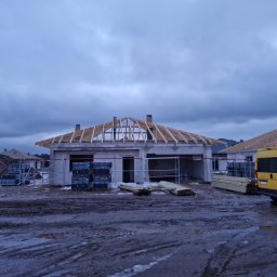 Budowa domu w trakcie krycia dachu, widoczne krokwie i kominy. Na pierwszym planie materiały budowlane i żółty samochód dostawczy na błotnistym terenie.