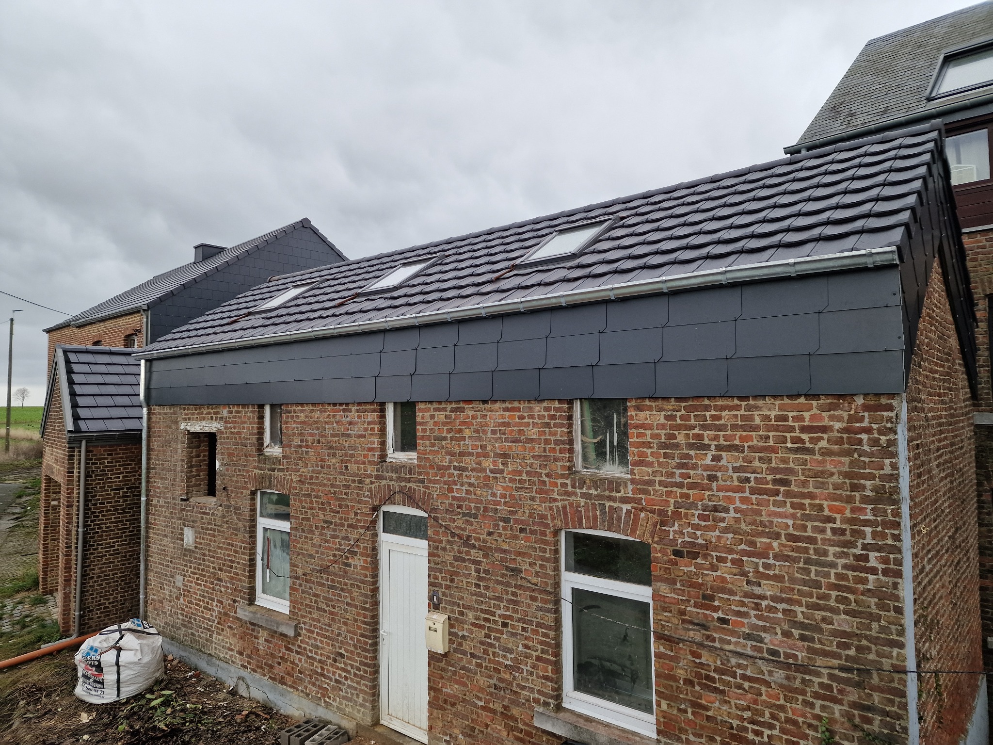 Ceglany budynek z nowym, ciemnoszarym dachem z oknami dachowymi i orynnowaniem. Widoczny fragment elewacji i worki z materiałami budowlanymi.