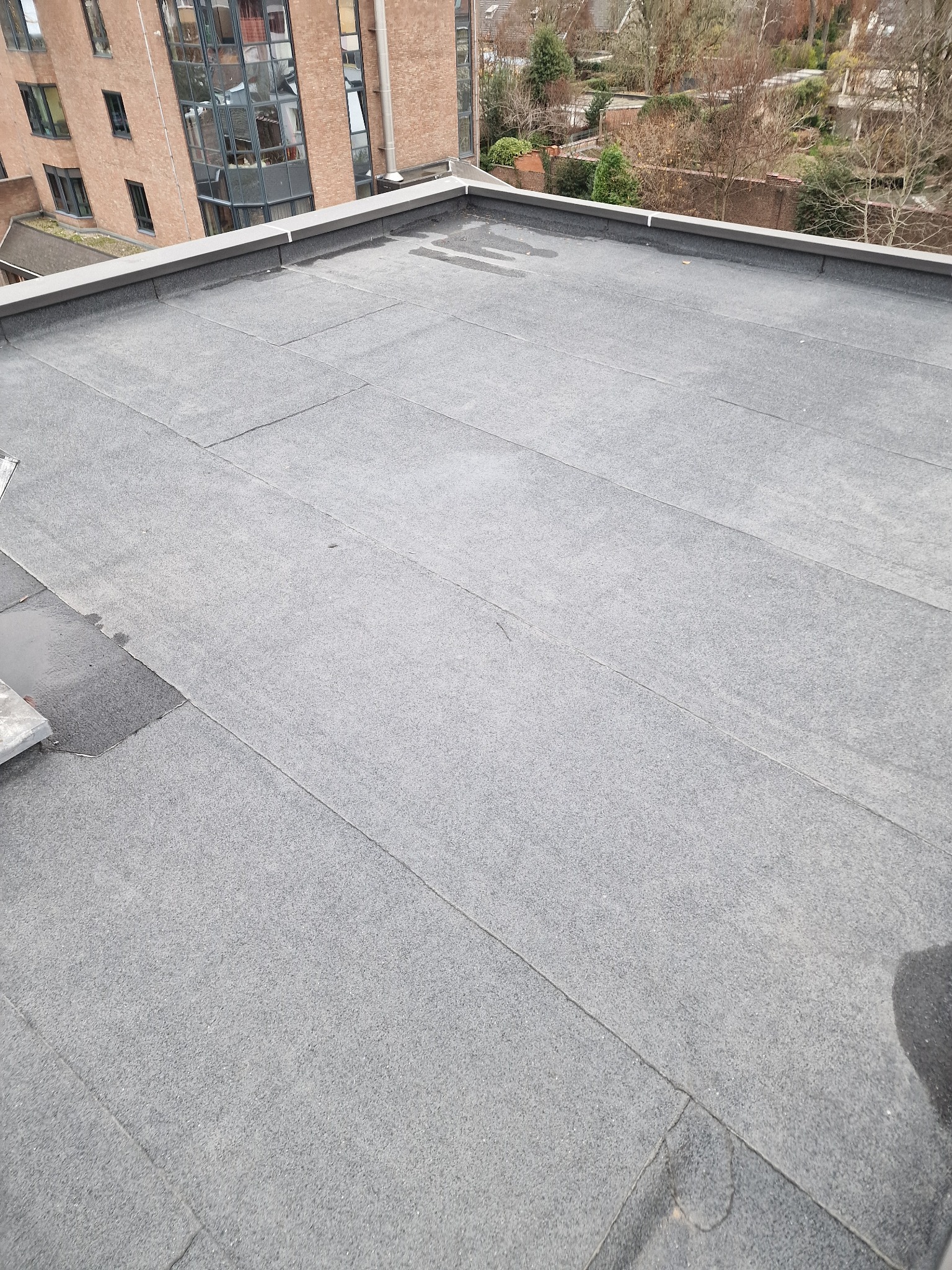 Szary, płaski dach pokryty papą termozgrzewalną, widok z góry. Na dachu widoczne ślady po wodzie. W tle budynki mieszkalne i drzewa.