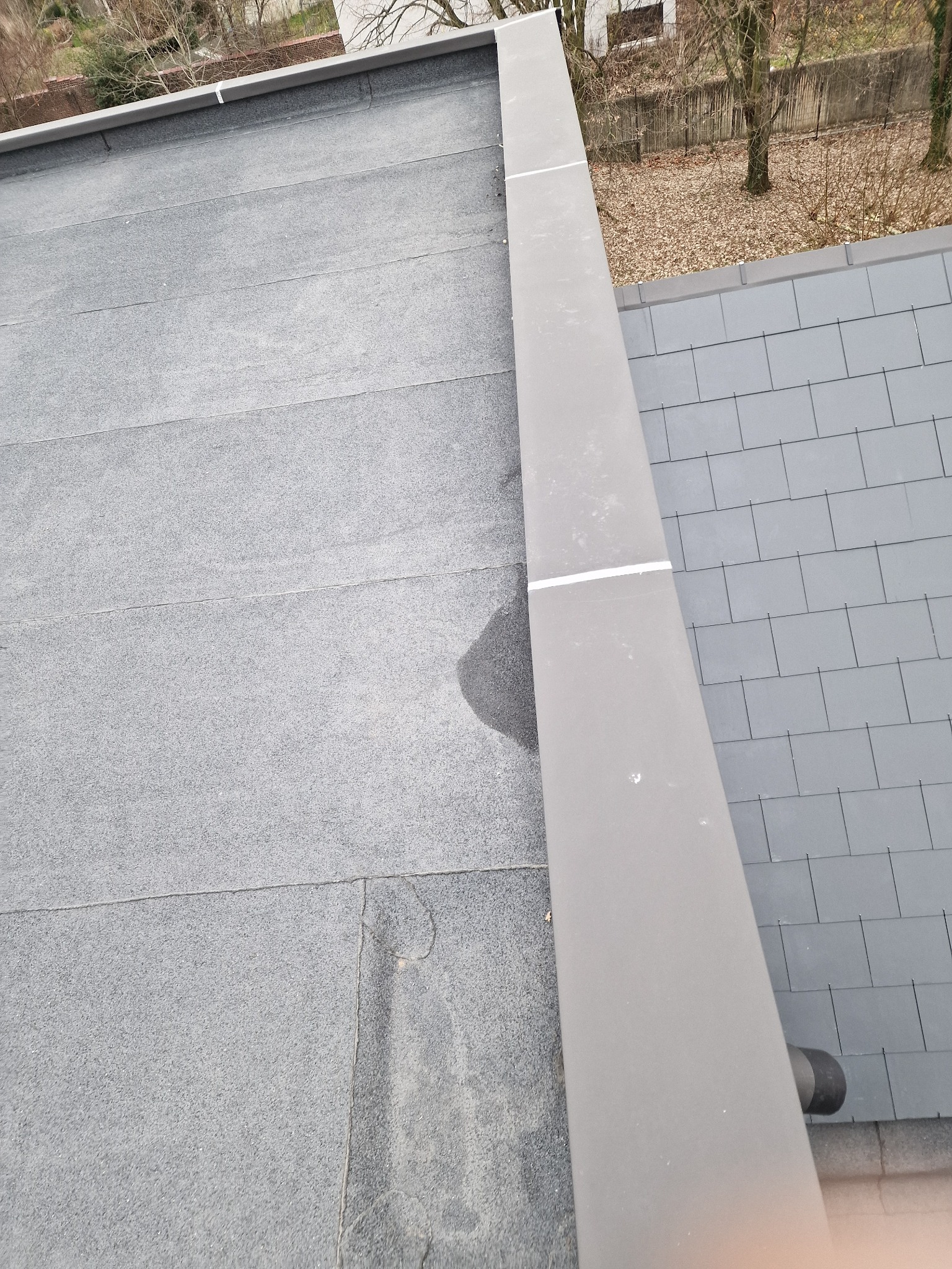 Szary, płaski dach pokryty papą, z widocznym fragmentem dachu z szarych dachówek i metalową obróbką blacharską. Widok z góry na gotowy dach.